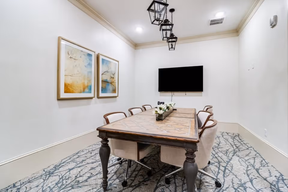 A modern conference room with a long wooden table surrounded by six beige swivel chairs. The room has white walls, two framed abstract paintings on one wall, a large flat-screen TV mounted on the opposite wall, and three black lantern-style pendant lights hanging from the ceiling. The floor is carpeted with a pattern resembling tree branches.
