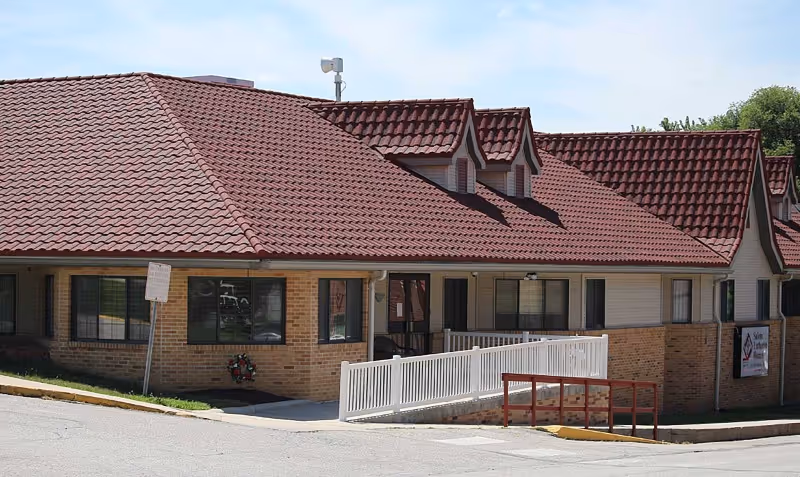 Exterior view of a single-story building with a red tiled roof and beige brick walls. The building has multiple windows and a white ramp leading to the entrance. There is a sign on the right side of the building indicating it is Salem Lutheran Homes and Rehabilitation Center.