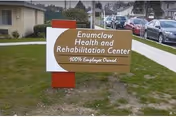 A sign for Enumclaw Health and Rehabilitation Center placed on a grassy area beside a sidewalk. The sign is brown and white with red supports and states that the center is 100% employee owned. In the background, there are parked cars and part of a building.