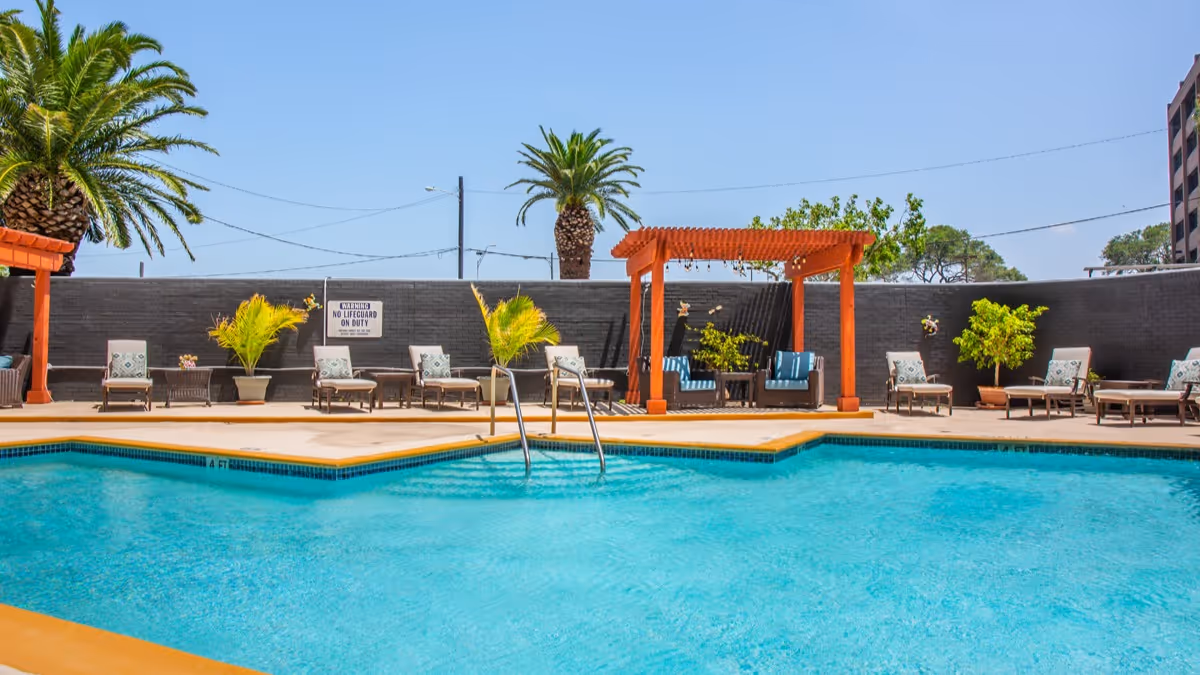 Outdoor swimming pool area with clear blue water, surrounded by lounge chairs with cushions, potted plants, and a wooden pergola with seating underneath. Palm trees and a clear blue sky are visible in the background.
