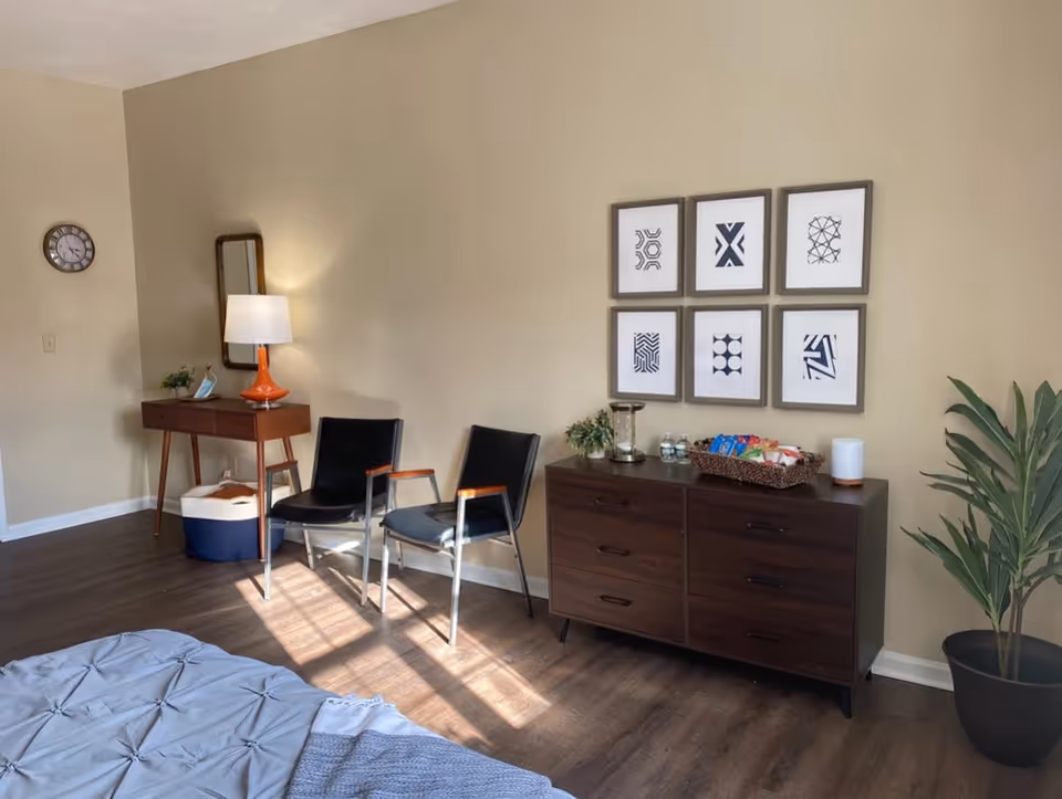 Sunlit bedroom area with a wooden dresser topped with decor and snacks, two chairs, a small writing table with a lamp and mirror, framed wall art, and a potted plant.