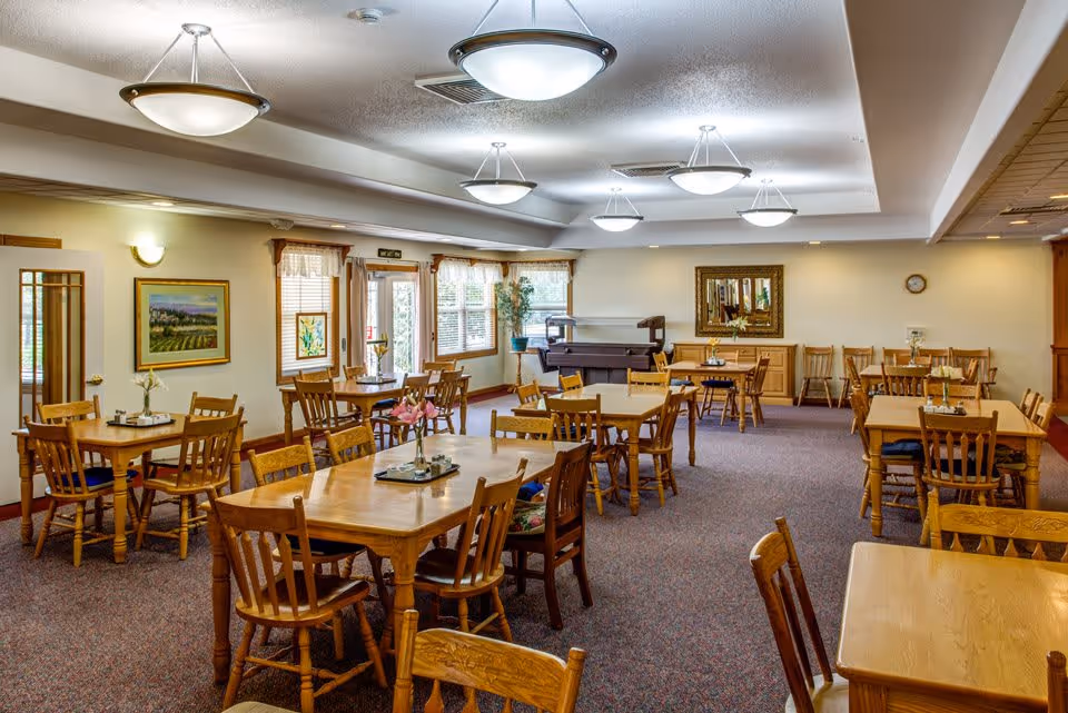 A spacious dining room with multiple wooden tables and chairs arranged neatly. The room is well-lit with ceiling lights and natural light coming through windows with curtains. There are framed paintings on the walls and a piano in the corner. The carpet is patterned and the overall atmosphere is warm and inviting.