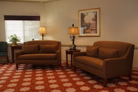 A cozy living room area with two brown sofas facing each other, a small round wooden table with a decorative lamp between them, a framed picture on the wall, a window with blinds, and a patterned red carpet covering the floor.