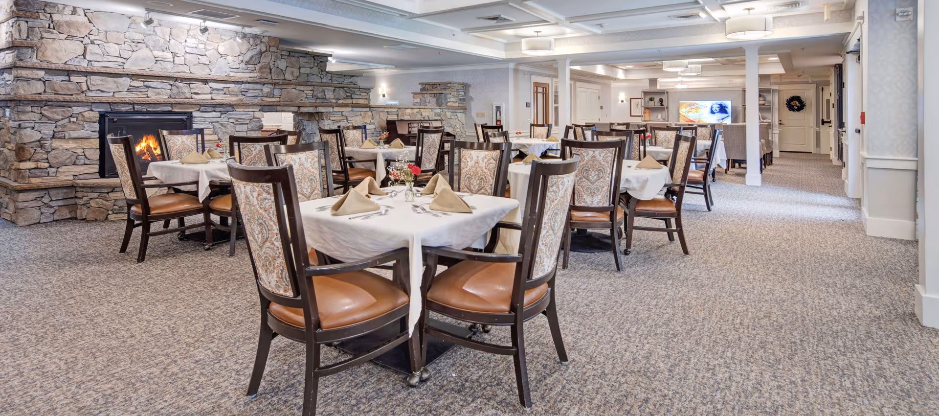 Spacious dining room with multiple set tables and upholstered chairs, a stone fireplace, and carpeted floor.
