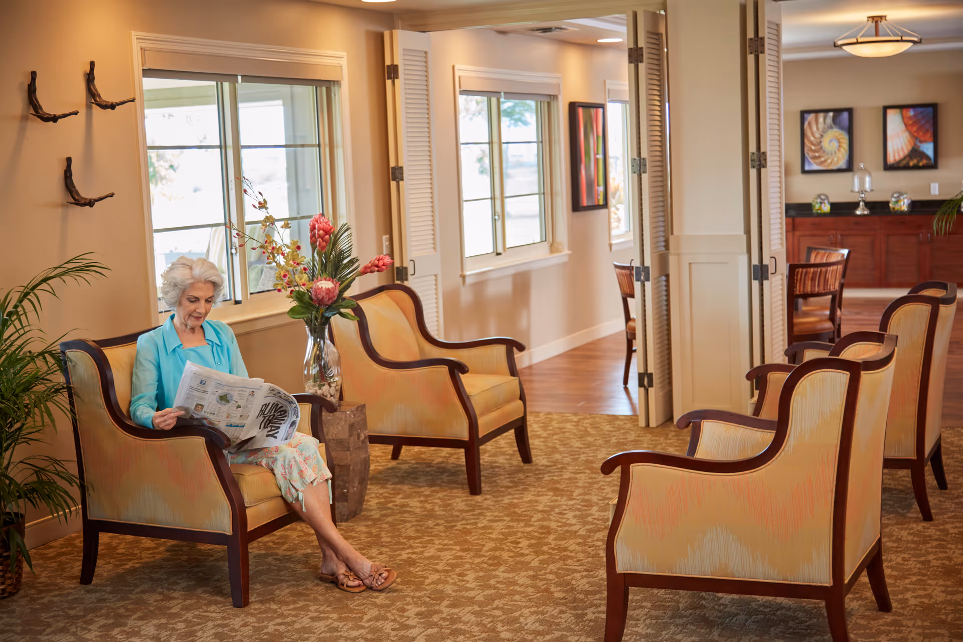 An elderly woman with white hair sits on a cushioned armchair reading a newspaper in a well-lit lounge area. The room features several similar armchairs, a large vase with flowers, windows letting in natural light, and framed artwork on the walls. The carpet has a patterned design and there are folding doors partially open to another room with wooden chairs and a sideboard.