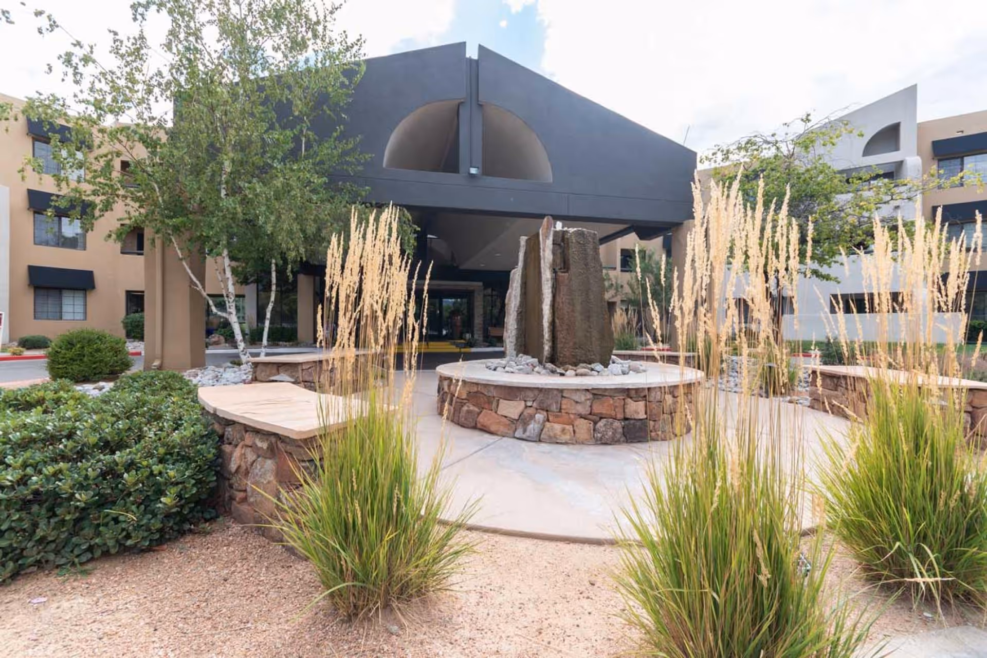 Front entrance of a senior living building with a stone fountain, tall grasses, and a covered porte-cochere.