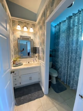 A bathroom with a white vanity and sink, a decorative mirror above the sink, and three light fixtures mounted on the wall. The walls have patterned wallpaper. To the right, there is a toilet and a shower area with a blue patterned shower curtain. The floor is tiled and there are dark gray bath mats in front of the sink and toilet.