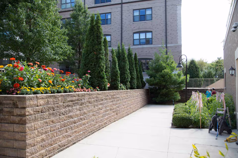 Concrete courtyard walkway with a raised brick planter filled with flowers and evergreens beside a multi-story building.