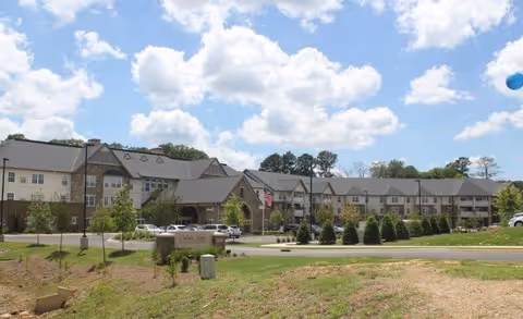 Exterior view of Cahaba Ridge Retirement Community, a large multi-story building with a combination of stone and siding facade, surrounded by landscaped greenery and a clear blue sky with scattered clouds.