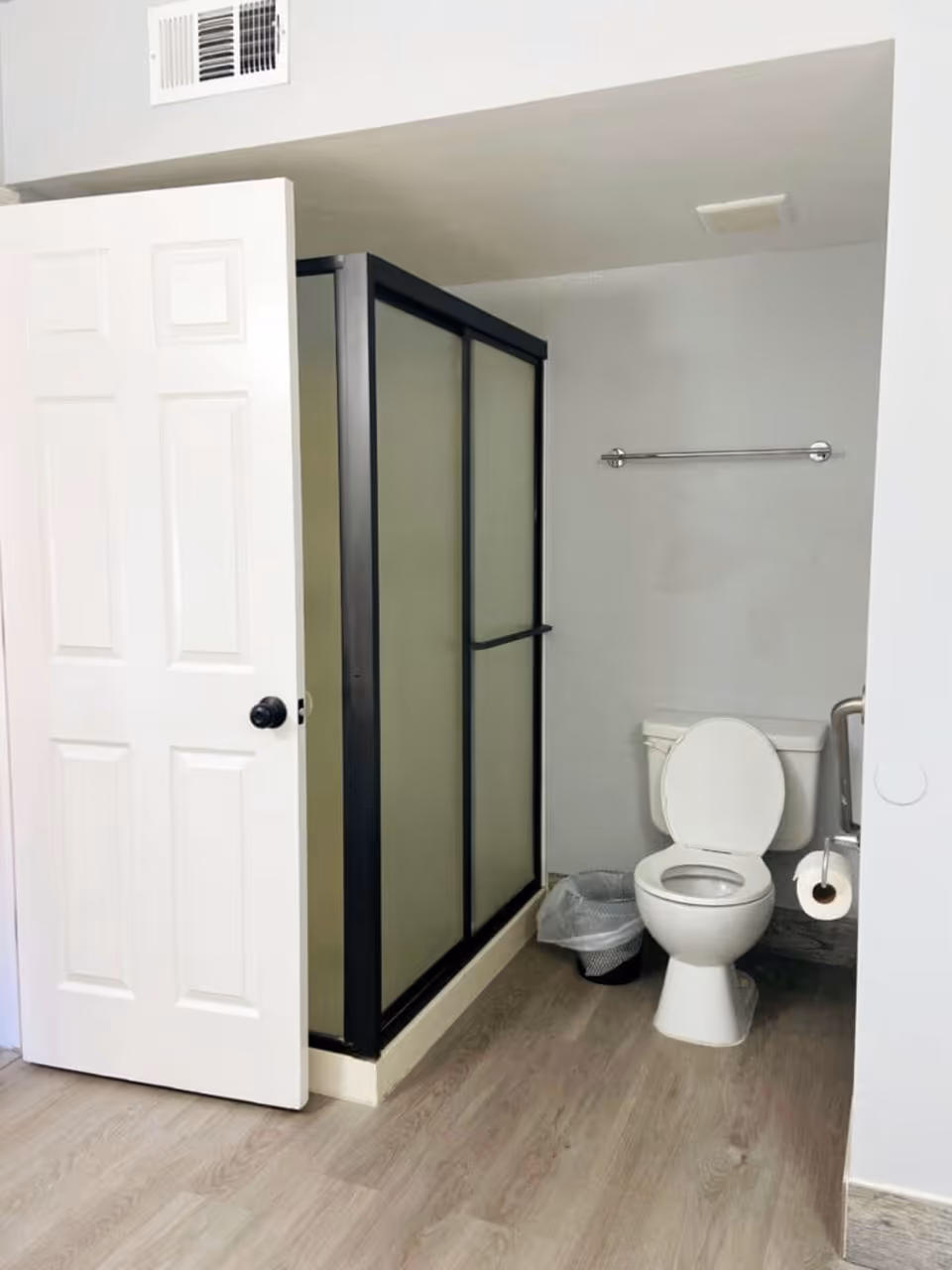 Bathroom featuring a frosted-glass shower stall, a toilet with open lid, a towel bar, and a partially open door.