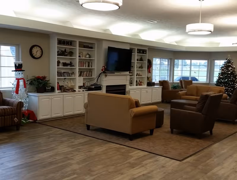 A cozy senior living common area with beige and brown armchairs and sofas arranged around a central rug. There is a white built-in shelving unit with a mounted flat-screen TV above a fireplace. The shelves hold books, decorations, and plants. A decorated Christmas tree and a lighted snowman figure are visible near the windows, which let in natural light. The room has wood flooring and modern ceiling lights.