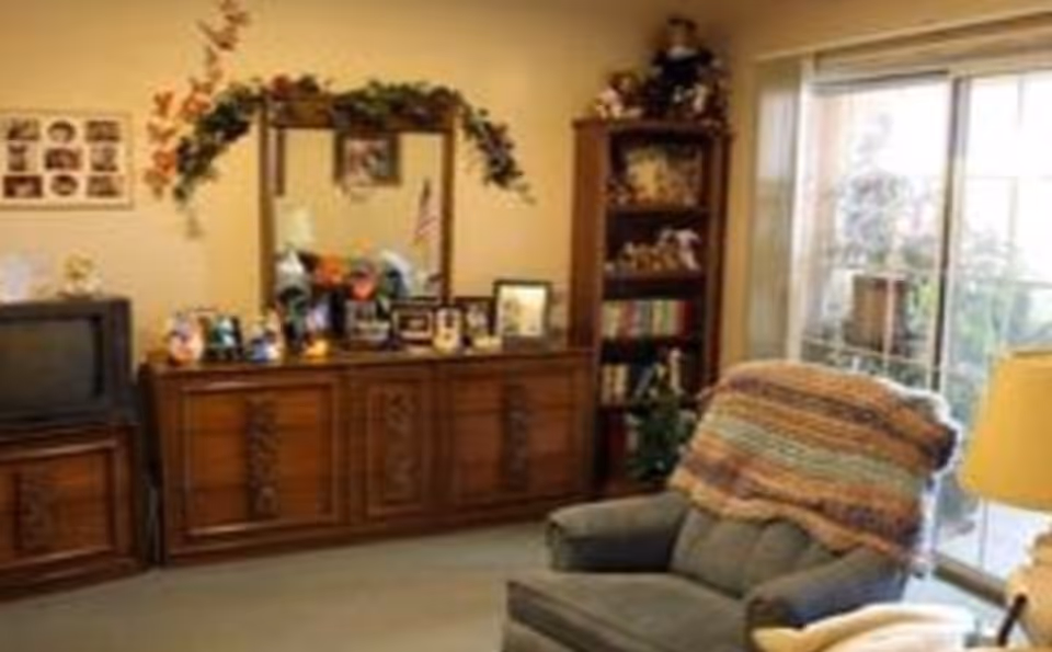 Cozy living room with an upholstered armchair draped with a blanket, wooden sideboard and mirror with photos and decorations, a bookshelf, TV, and sliding glass door.