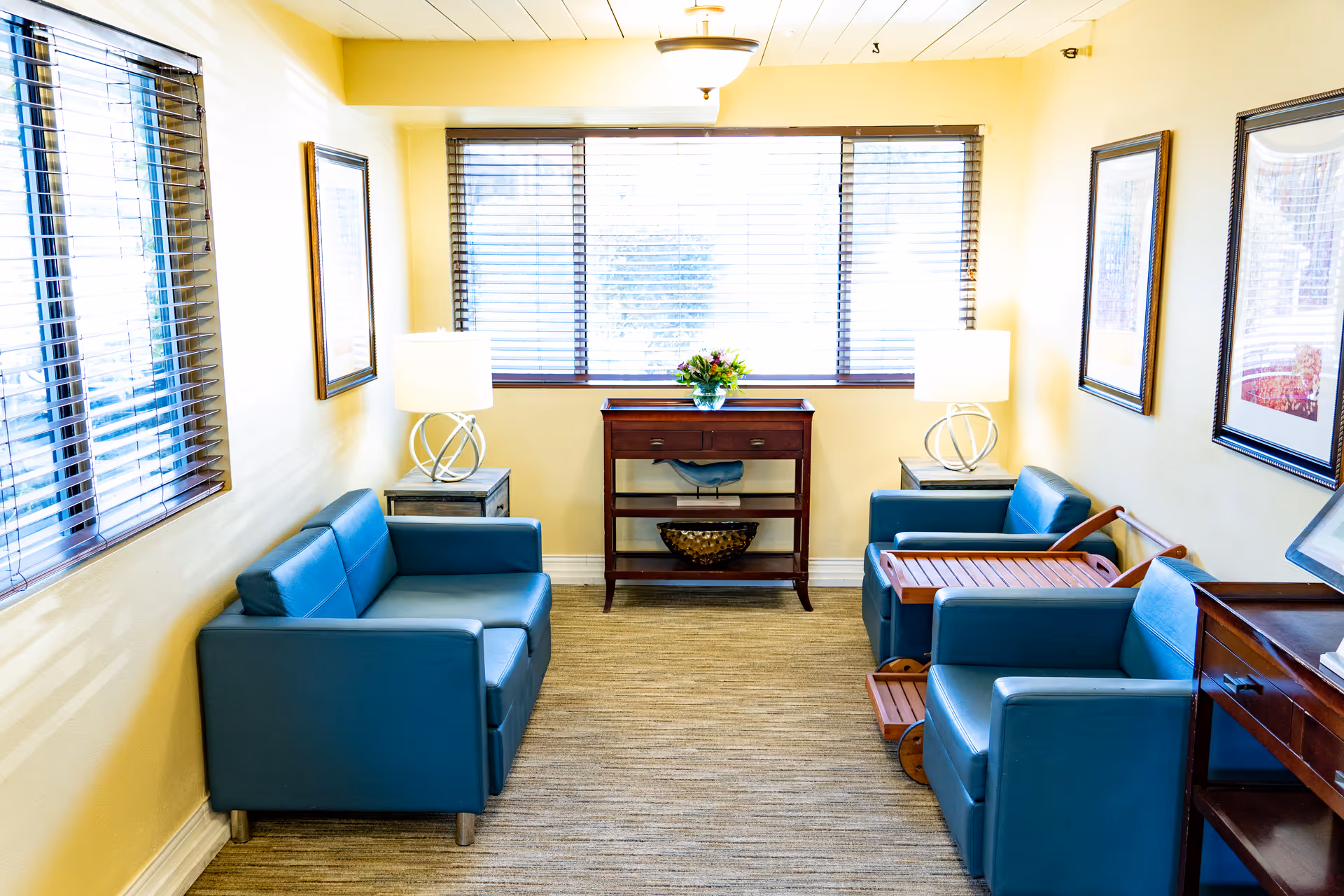 A bright, cozy sitting area with two blue armchairs and a blue loveseat arranged around a small wooden side table with a lamp on each side. A wooden console table with a flower arrangement is placed against the wall under a large window with blinds. The walls are painted light yellow and decorated with framed artwork.
