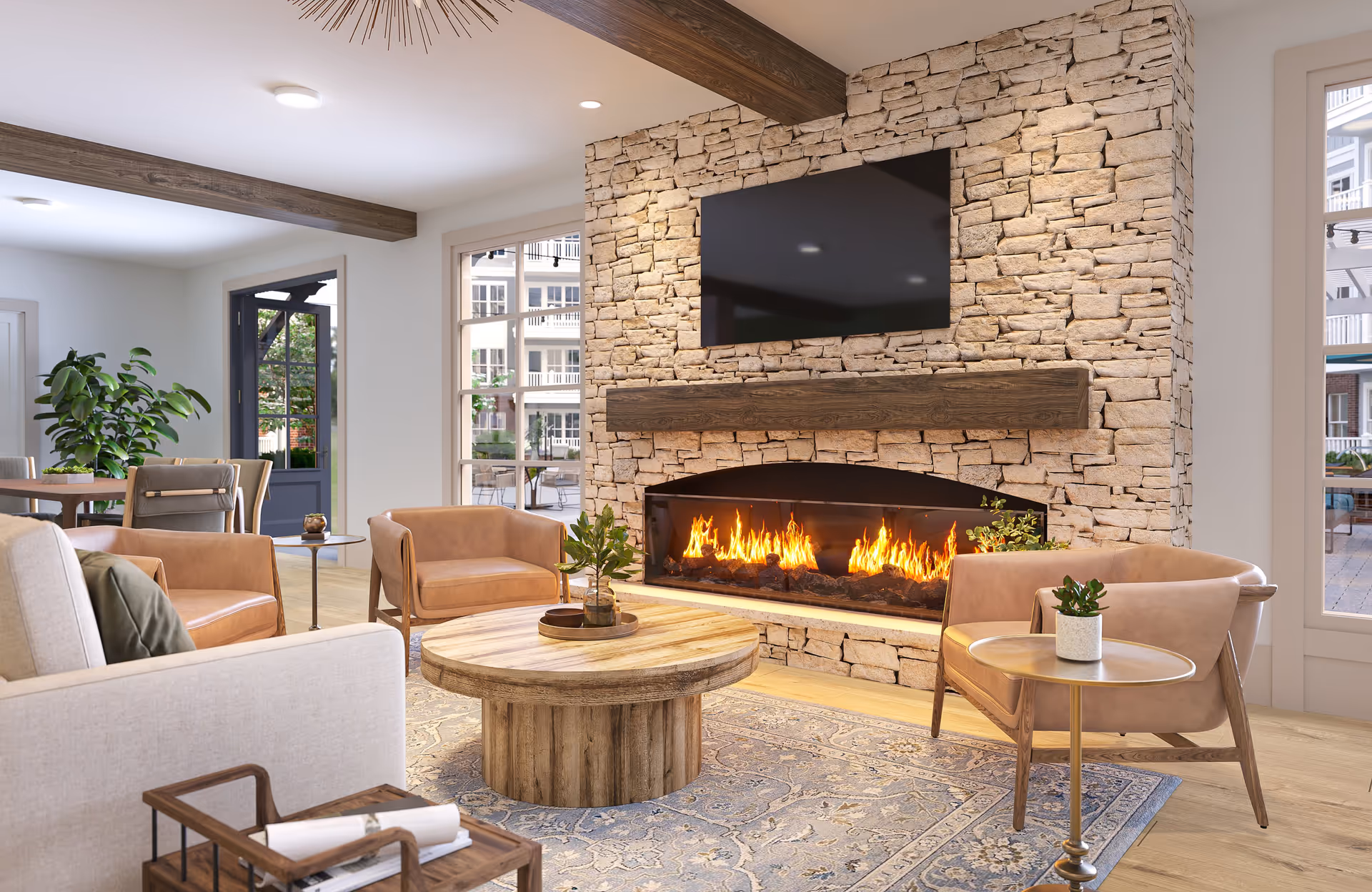A cozy living room area featuring a stone fireplace with a modern fire burning inside. Above the fireplace is a large flat-screen TV mounted on the stone wall. The room has wooden beams on the ceiling, light-colored walls, and large windows letting in natural light. There are several comfortable chairs and a round wooden coffee table with small plants on it. A patterned rug covers the wooden floor, and a door opens to an outdoor patio area.
