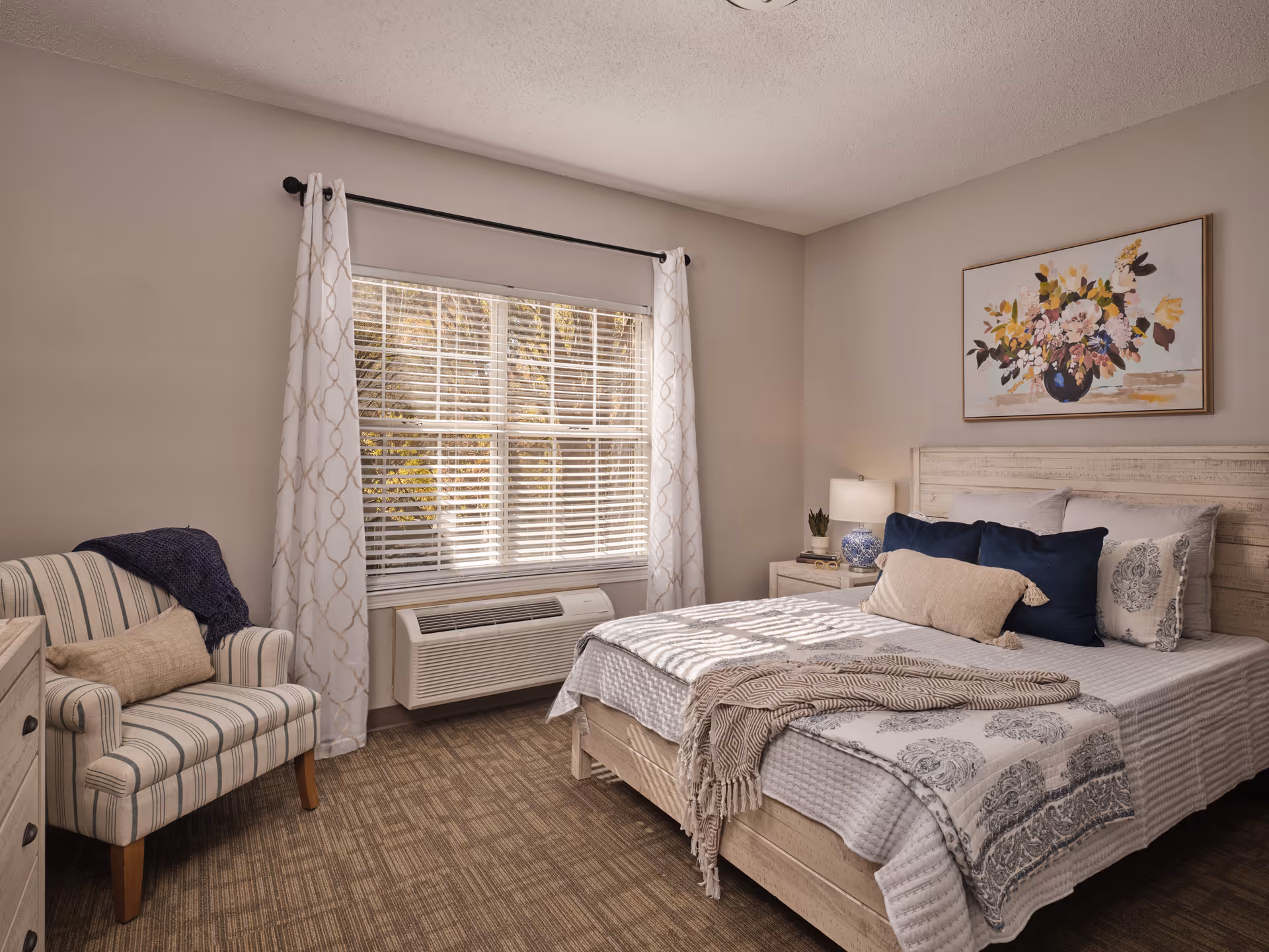 A cozy bedroom with a large window covered by white curtains and blinds. The room features a bed with white and patterned bedding, several pillows, and a beige throw blanket. Next to the bed is a nightstand with a lamp and a small plant. A striped armchair with a beige pillow and a dark blue throw blanket is positioned near the window. A framed floral painting hangs on the wall above the bed. The floor is carpeted in a neutral tone.