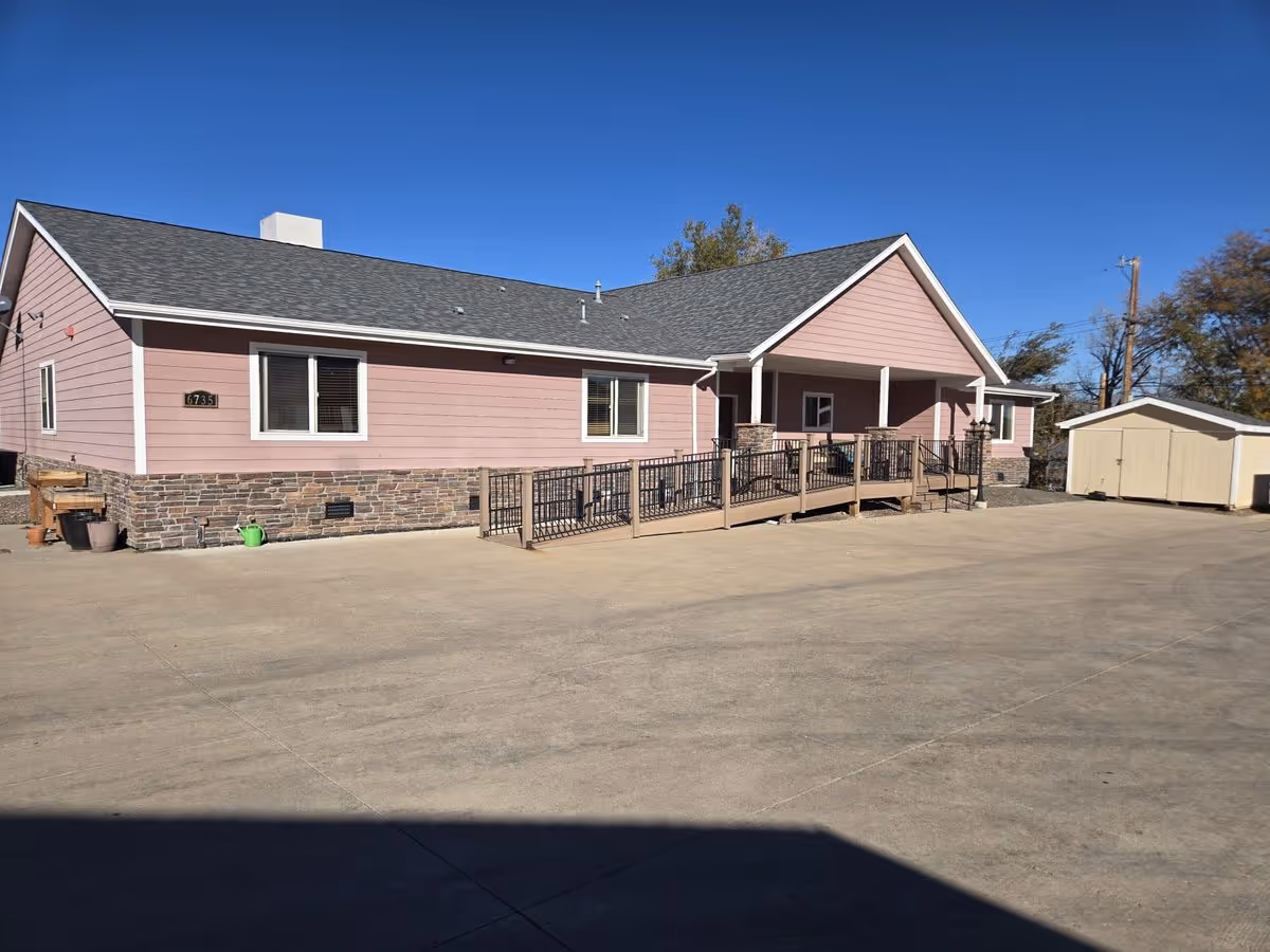 Single-story pink building with a ramped front porch and stone foundation beside a storage shed under a clear blue sky.