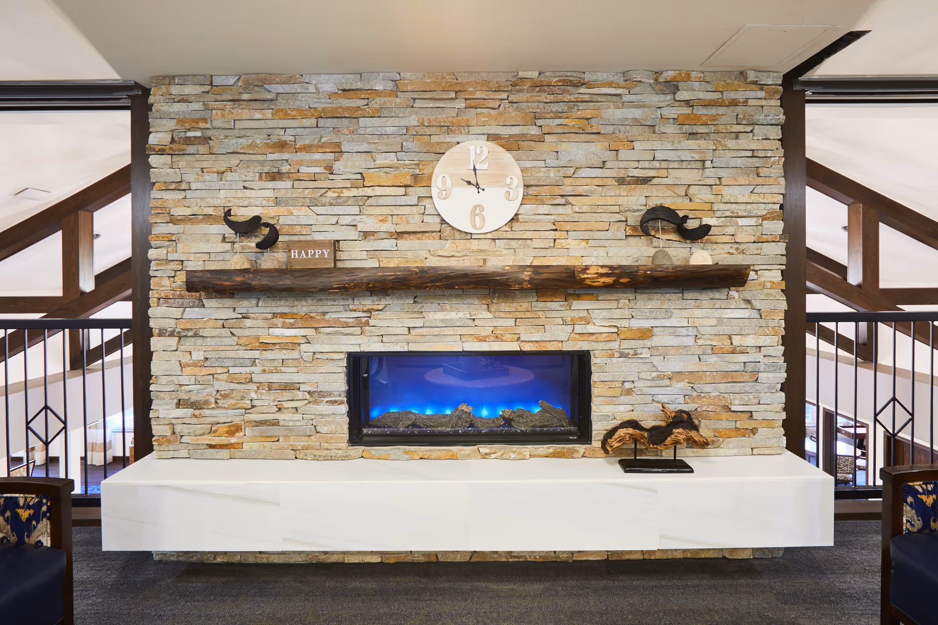 A stone wall with a mounted electric fireplace emitting blue light. Above the fireplace is a rustic wooden mantel with decorative items including a clock, a small sign that says 'HAPPY', and bird figurines. The area is part of an interior space with visible wooden beams and railings on either side.
