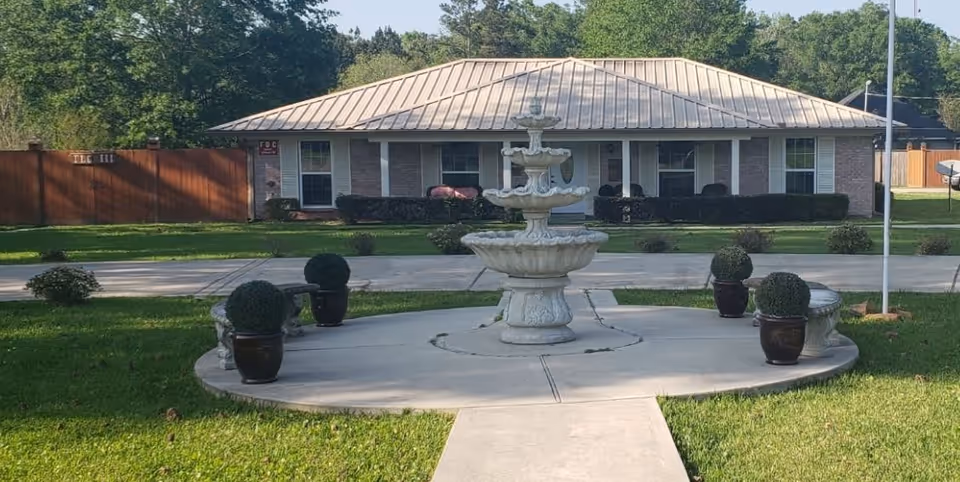 A single-story brick building with a metal roof is visible behind a circular concrete area featuring a multi-tiered white fountain in the center. The circular area is surrounded by four potted plants and two curved stone benches. The scene is set on a grassy lawn with trees in the background under a clear sky.
