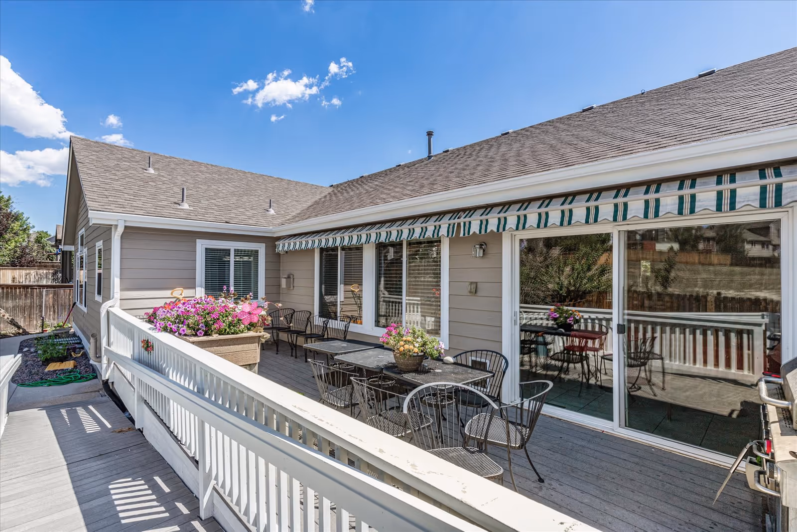Outdoor patio area at a senior living facility with a wooden deck, white railing, several black metal tables and chairs, flower pots with colorful flowers, and a striped green and white awning above the sliding glass doors leading inside.