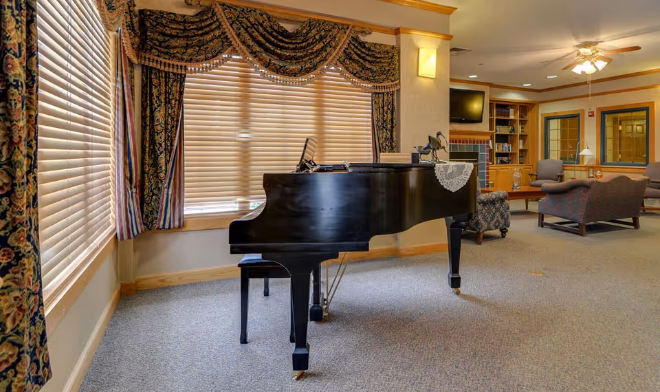 A spacious common area with a black grand piano in the foreground, large windows with patterned curtains and blinds, and comfortable seating including armchairs and a loveseat. There is a built-in bookshelf, a wall-mounted TV, and a ceiling fan with lights in the background.