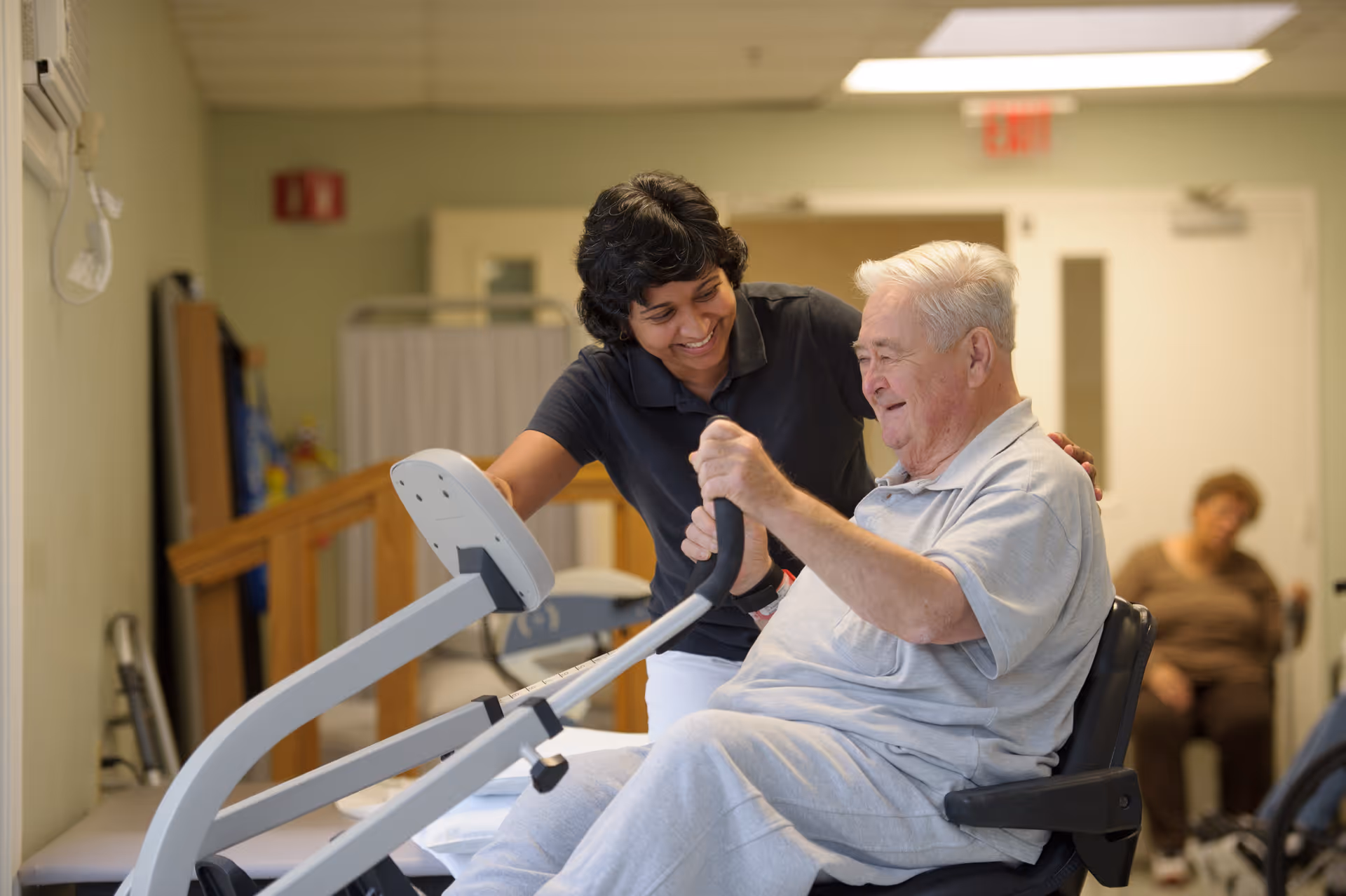 A senior man using a seated exercise machine with the assistance of a smiling female caregiver in a health center room. Another person is seated in the background.