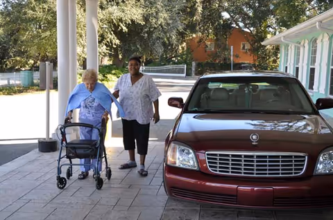 An elderly woman using a walker is being assisted by a caregiver outside near a maroon car parked by a building with white pillars and green window shutters. Trees and a fence are visible in the background.