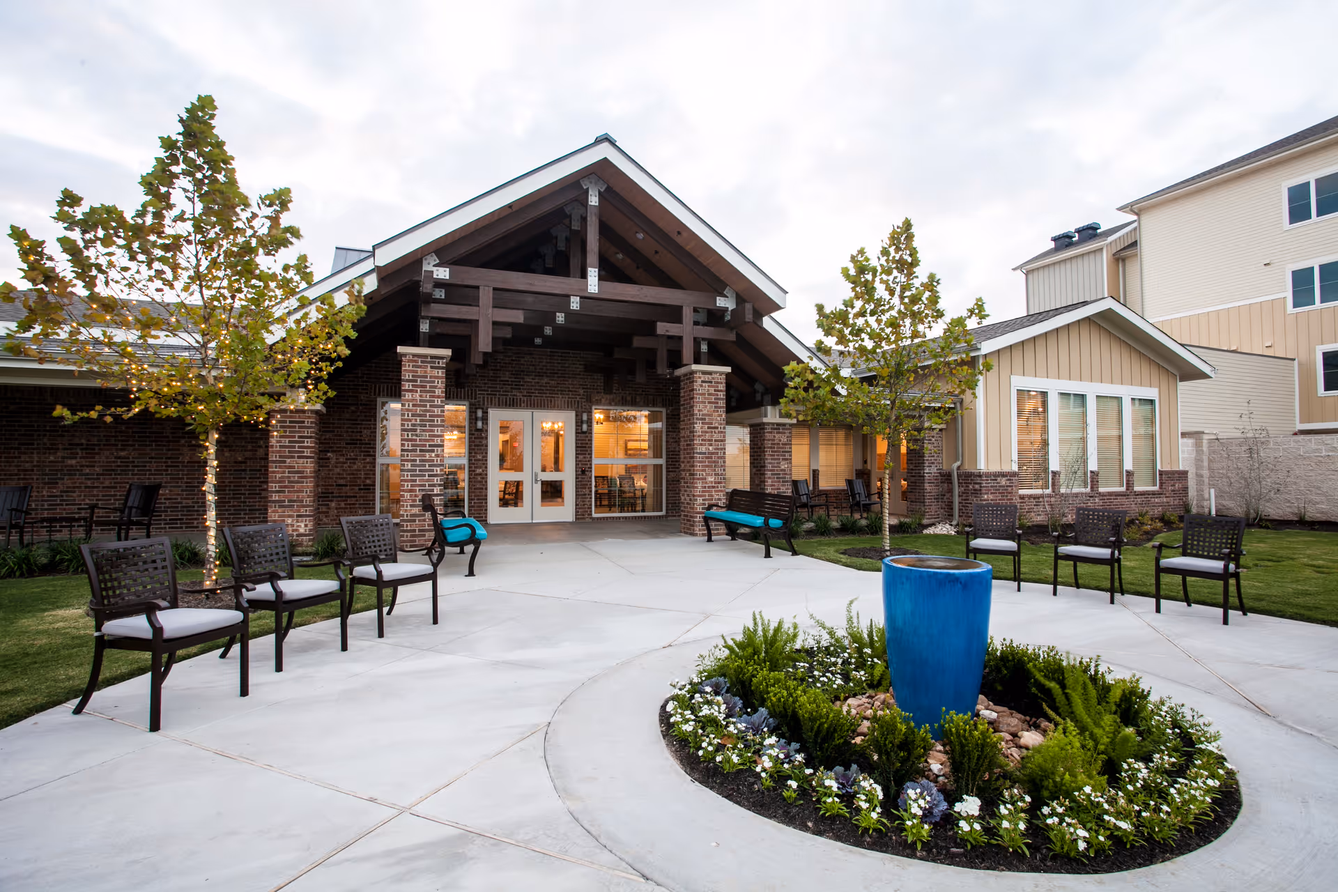 Outdoor view of the entrance to The Delaney at Lake Waco facility featuring a covered porch with brick pillars, several chairs and benches arranged on a concrete patio, small trees, and a circular flower bed with a large blue ceramic planter in the center.