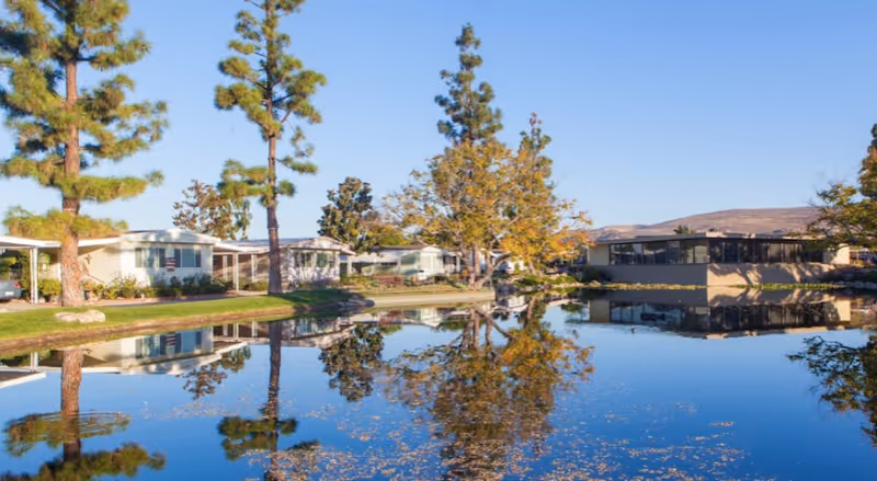 A serene outdoor scene at Lake Park Yorba Linda featuring a calm pond reflecting nearby trees and single-story buildings under a clear blue sky.