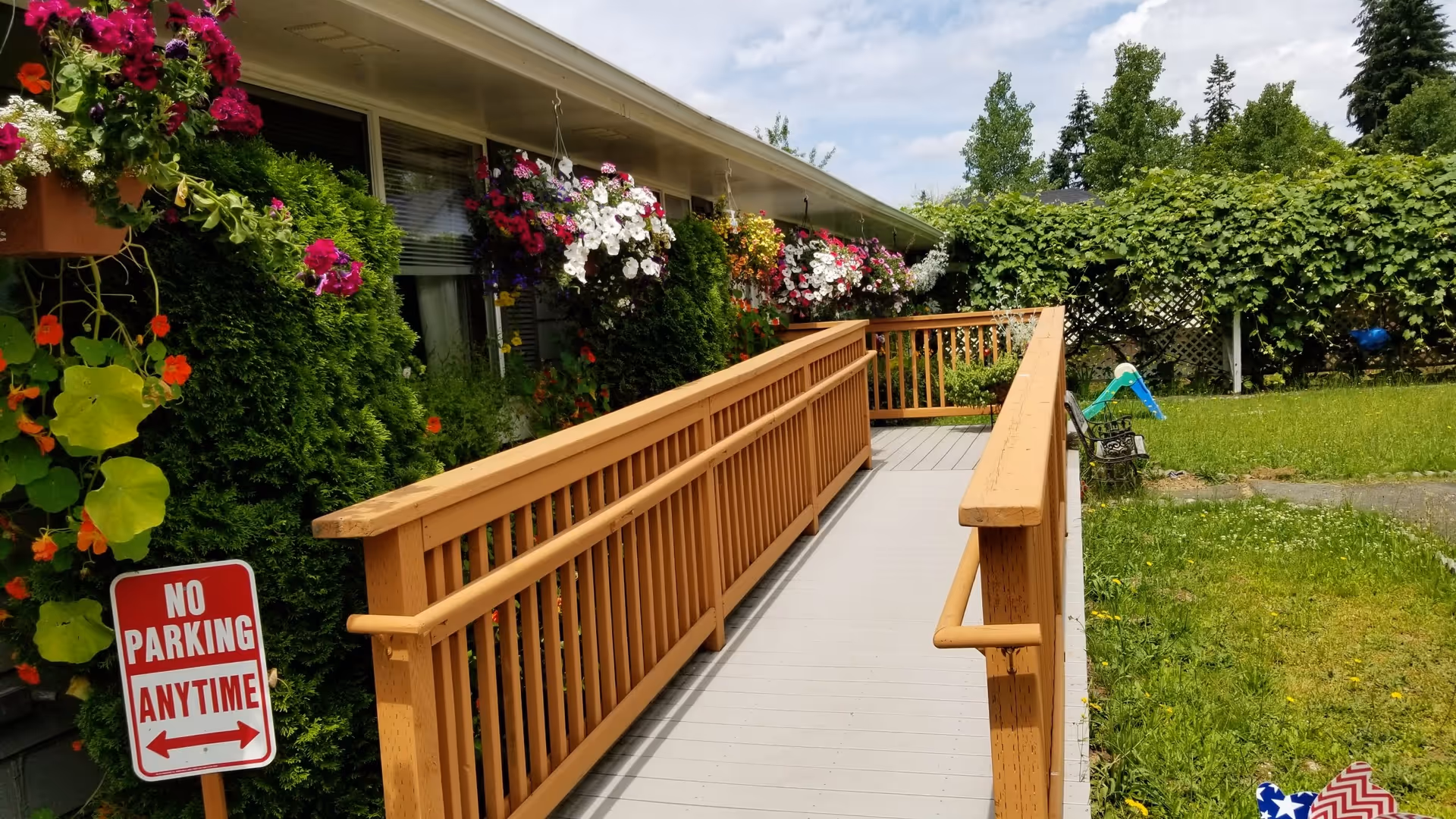 Outdoor view of a wooden wheelchair ramp leading to the entrance of a building. The ramp is surrounded by green bushes and colorful hanging flower baskets. A 'No Parking Anytime' sign is visible on the left side near the ramp. The sky is partly cloudy.