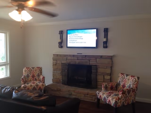 A cozy living room with a stone fireplace in the center, a flat-screen TV mounted above it, two patterned armchairs on either side of the fireplace, and a dark leather sofa in the foreground. A ceiling fan with lights is visible on the ceiling, and a window is on the left wall.