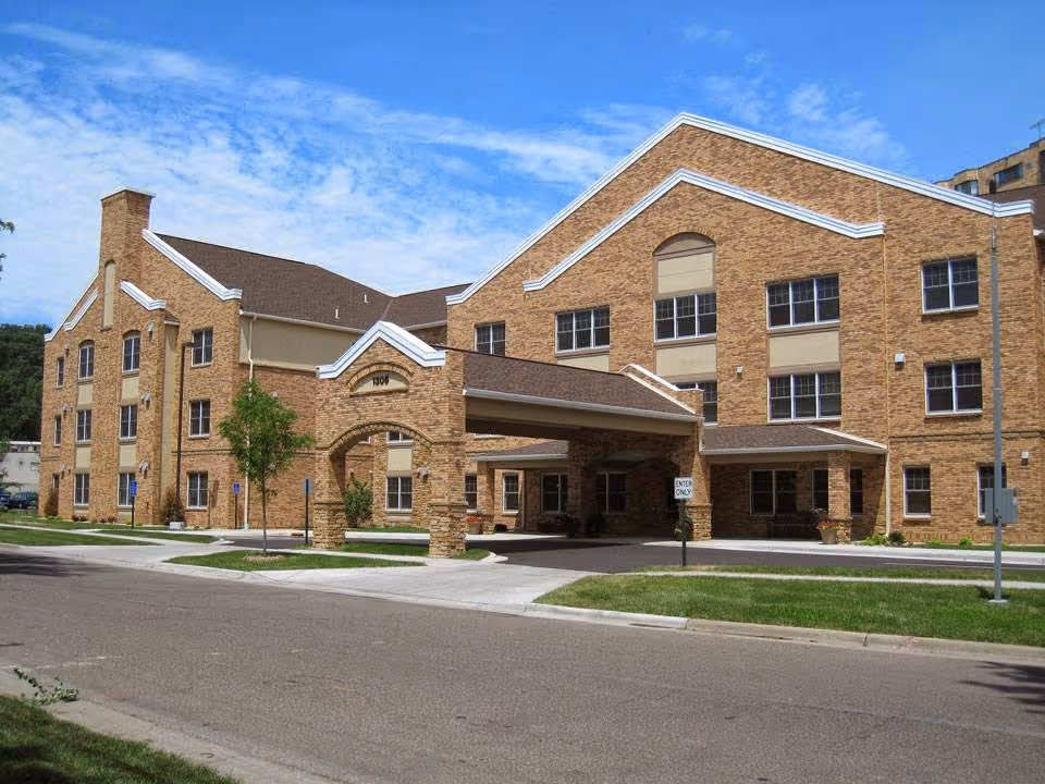 Exterior view of a large brick senior living facility building with multiple windows and a covered entrance driveway under a blue sky with scattered clouds.