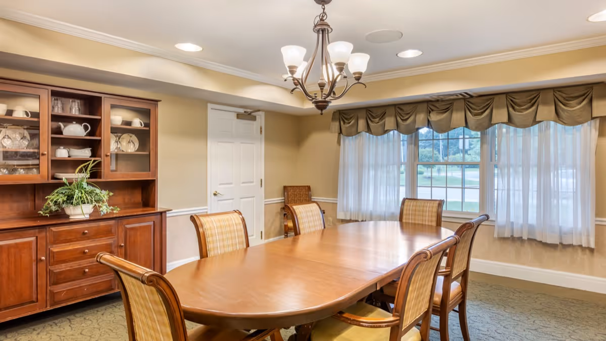 A dining room with a large wooden table surrounded by six wooden chairs with plaid upholstery. There is a wooden cabinet with glass doors displaying dishes and glassware on the left side. A chandelier with five lights hangs above the table. A window with sheer white curtains and a valance lets in natural light.