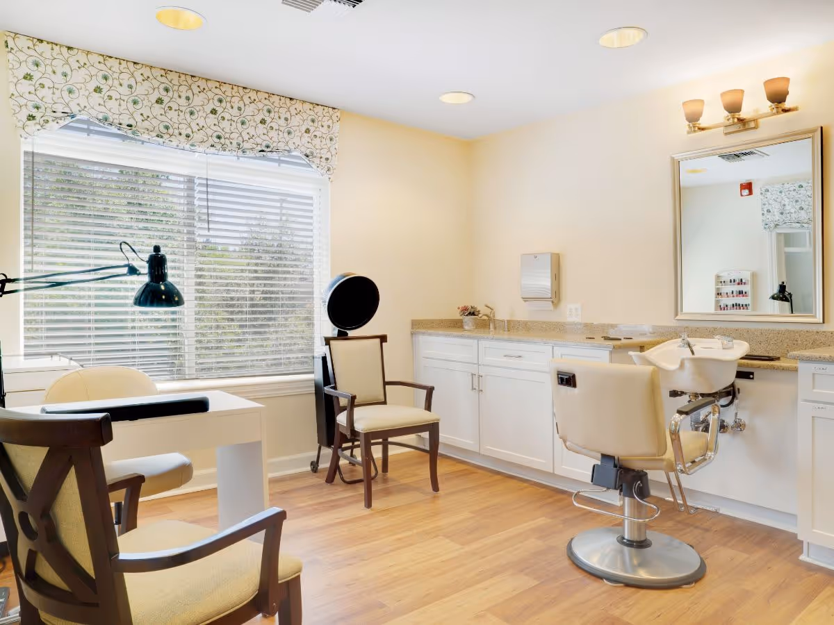 Bright salon-style room with styling chairs, a wash basin, mirror, manicure table and a large window with blinds.