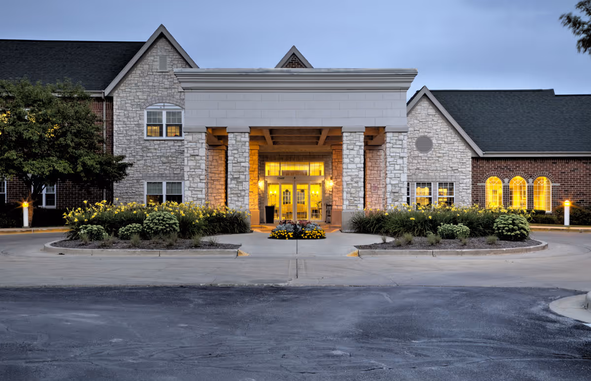 Exterior front view of a senior living facility building at dusk with stone and brick facade, illuminated entrance, landscaped flower beds, and a driveway in front.