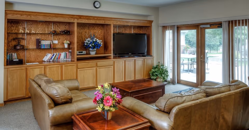 A cozy living room with two brown leather sofas facing a wooden entertainment center that holds a flat-screen TV, books, and decorative items. There is a wooden coffee table and a side table with a colorful flower arrangement. Large glass doors with vertical blinds open to an outdoor patio area with tables and chairs.
