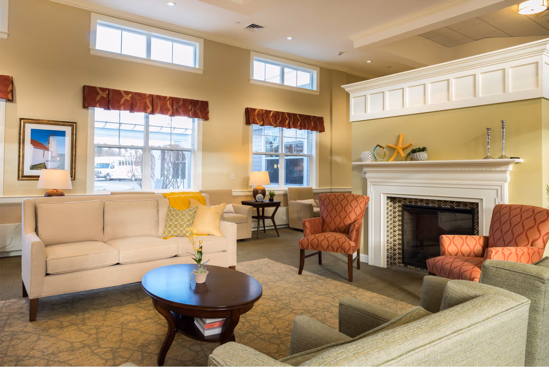 A cozy living room area with a beige sofa adorned with yellow and green pillows, two red patterned armchairs, and a green armchair. A dark wooden oval coffee table with a small potted plant sits in the center on a patterned carpet. The room features a white fireplace decorated with a starfish and other ornaments. Large windows with red valances allow natural light to fill the space, and there are two table lamps on side tables near the windows.