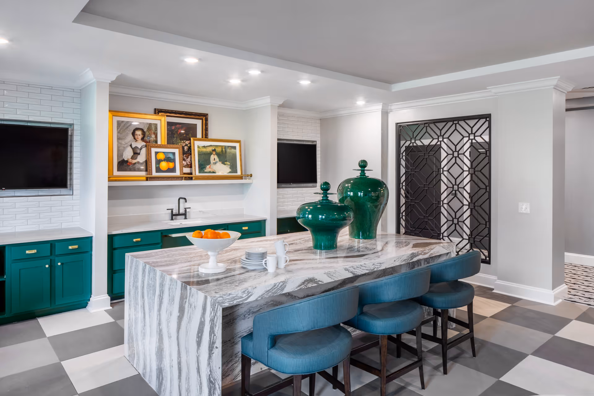 Modern interior space with a large marble island countertop surrounded by blue upholstered chairs. The countertop has two large green decorative vases and a white bowl with oranges. The background features green cabinetry with gold handles, two mounted flat-screen TVs, a small sink, and framed artwork displayed on a shelf. The floor has a checkered pattern in shades of gray and white, and a decorative black metal screen is visible on the right wall.