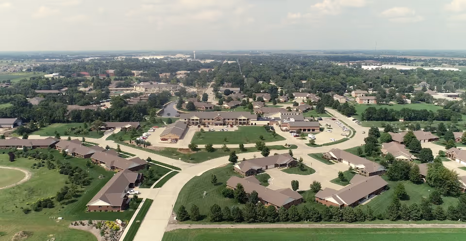 Aerial view of Schowalter Villa senior living facility showing multiple single-story buildings with brown roofs arranged around green lawns and trees, with roads and parking areas connecting the buildings. The surrounding area includes residential neighborhoods and open fields under a partly cloudy sky.