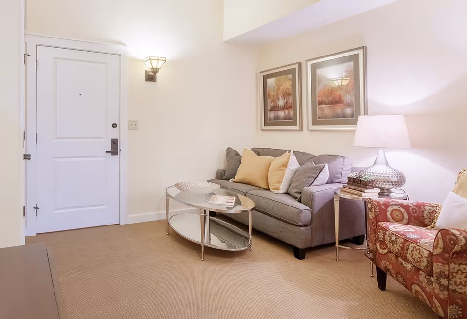 A cozy living room with a gray sofa, glass coffee table, patterned armchair, table lamp, wall art, and a white entrance door.