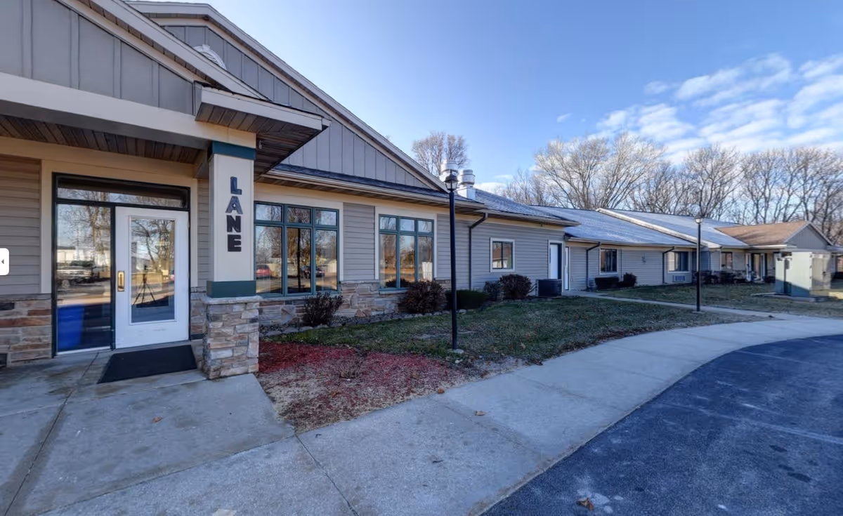 Exterior view of Cedar Vale Assisted Living facility showing a single-story building with beige siding, stone accents, multiple windows, a white door with glass panels, and a sidewalk curving around the front. There are lamp posts and leafless trees in the background under a partly cloudy sky.