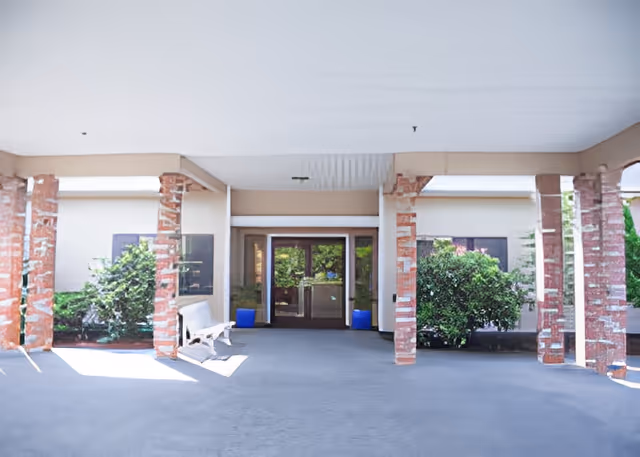 Covered entrance area of a building with brick pillars and glass double doors. There are green bushes on either side of the entrance and a white bench on the left side under the covered area.