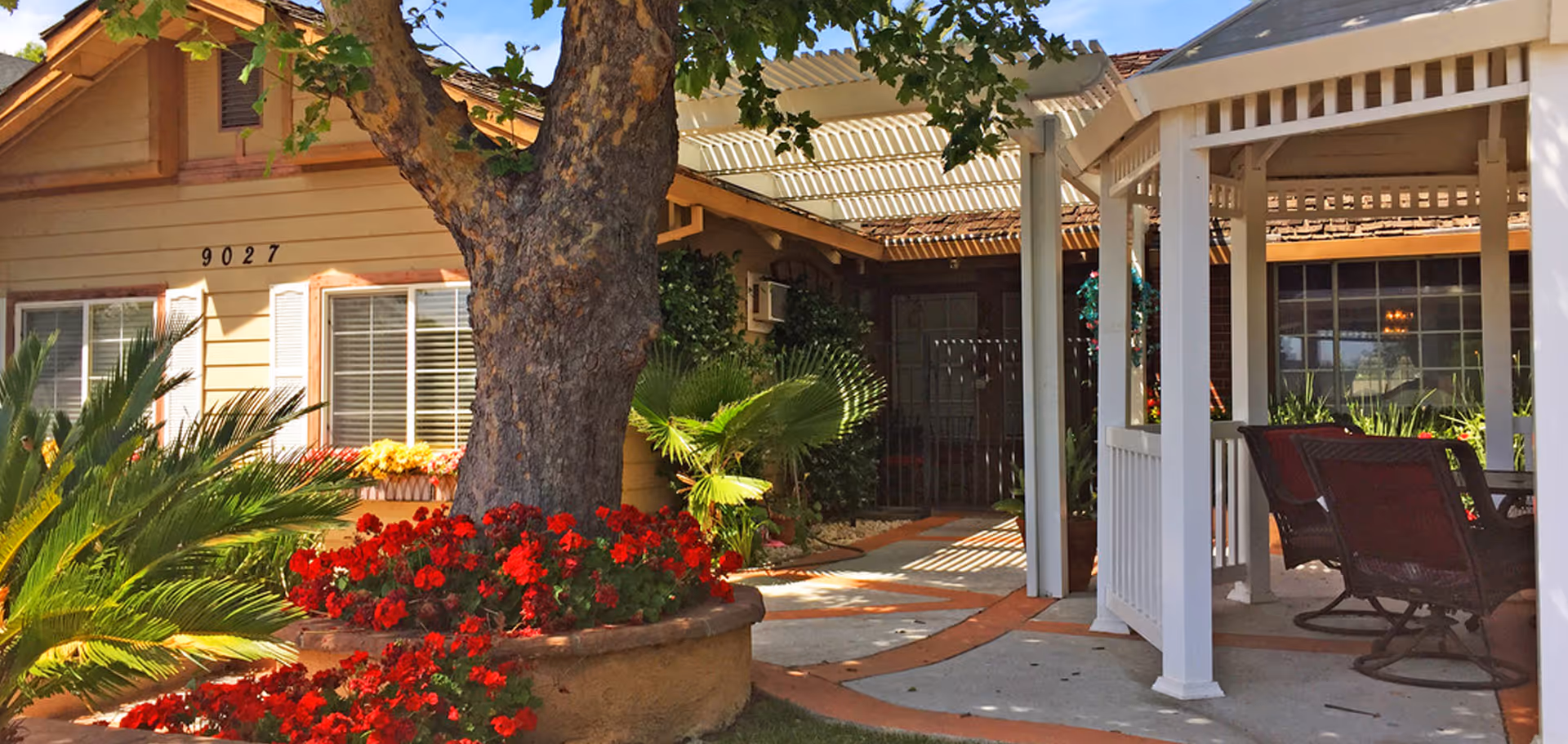 Outdoor garden area of a senior living facility with a large tree surrounded by red flowers, green plants, a beige building with windows and shutters, and a white gazebo with chairs underneath.