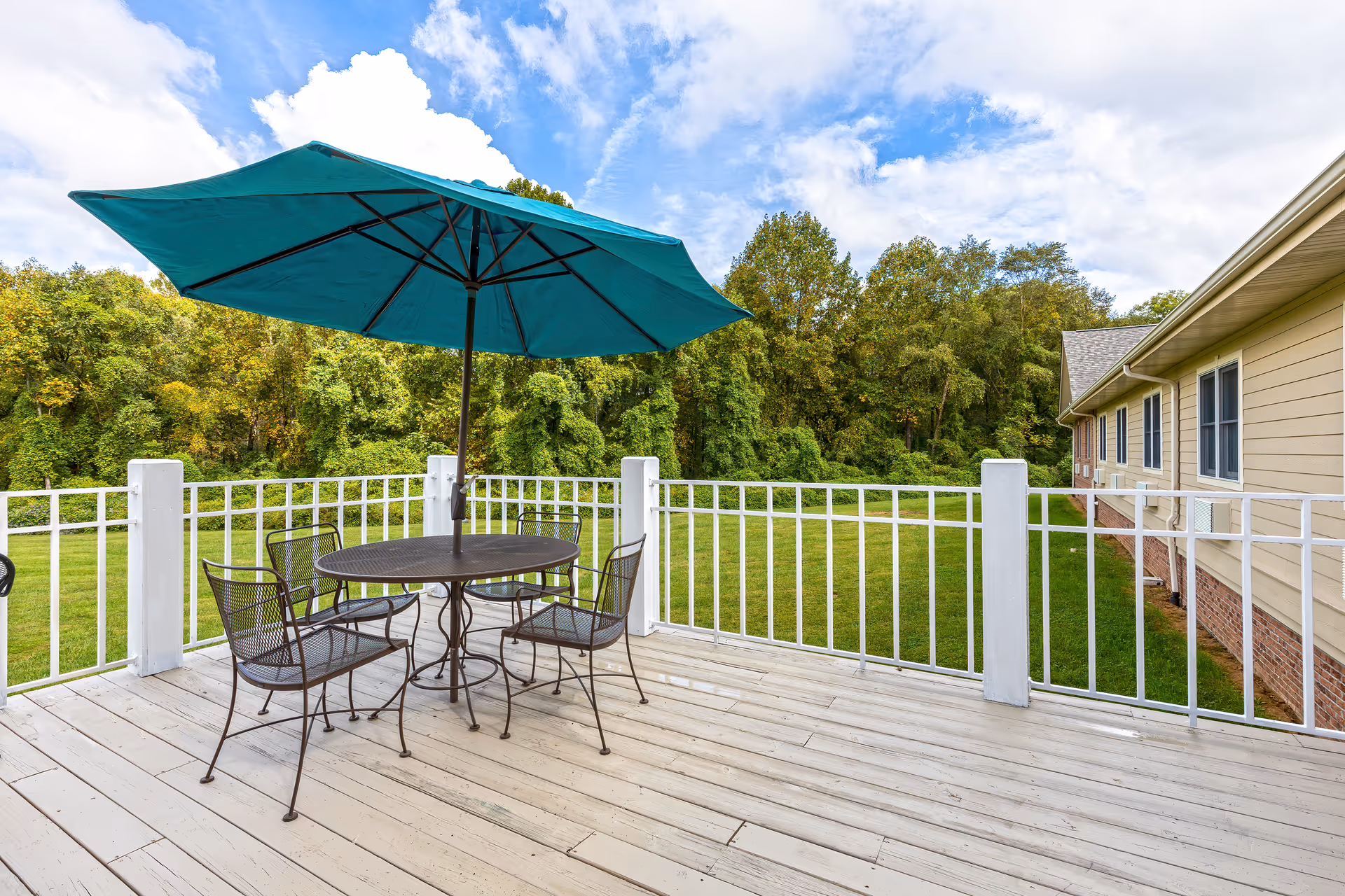 Outdoor patio area with a round metal table and four matching chairs under a large teal umbrella. The patio has white railings and overlooks a grassy lawn with trees in the background under a partly cloudy sky.