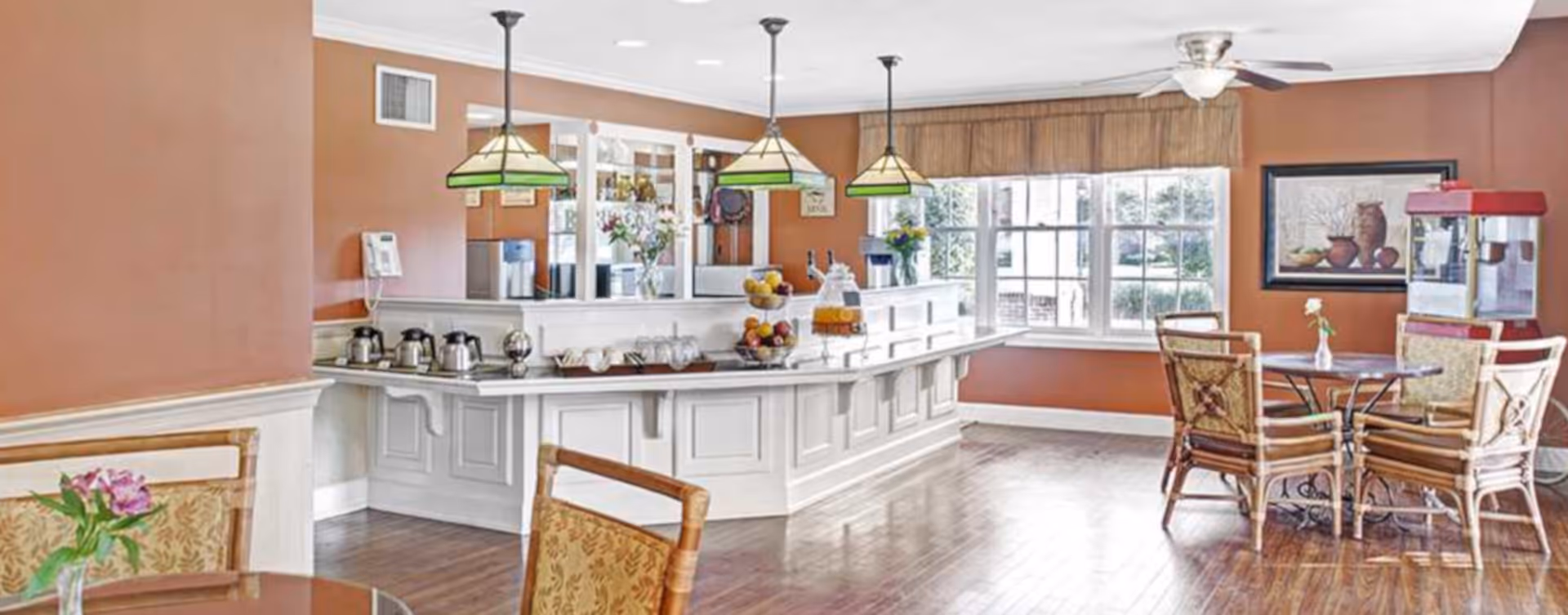 Bright communal dining area with a white serving counter, pendant lights, tables and chairs by a large window.