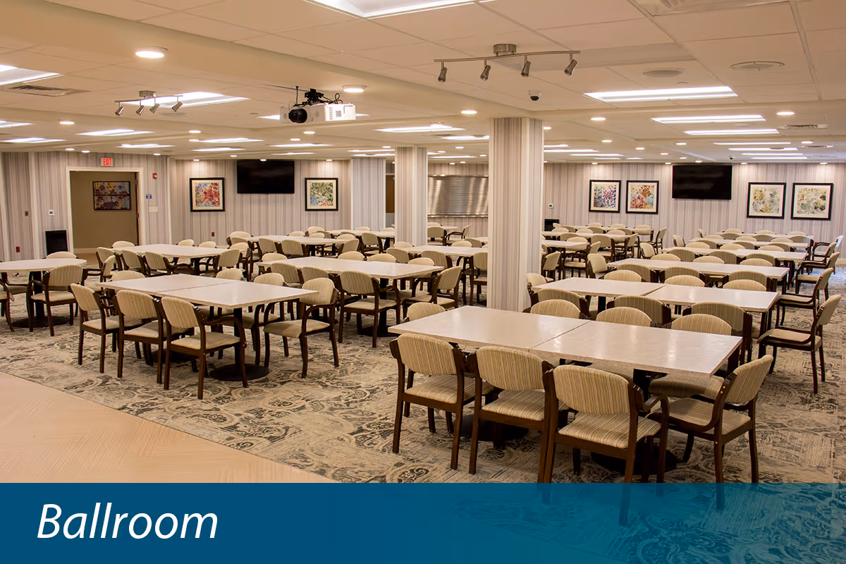 A large ballroom with multiple rectangular tables and beige cushioned chairs arranged neatly. The room has patterned carpet flooring, striped wallpaper, framed artwork on the walls, and ceiling lights. There are two large flat-screen TVs mounted on the walls and a projector hanging from the ceiling.