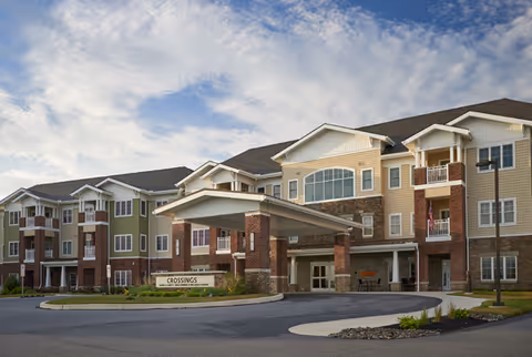 Exterior view of a multi-story senior living facility building with a covered entrance and a sign that reads 'Crossings'. The building features a combination of brick and siding with balconies and large windows under a partly cloudy sky.