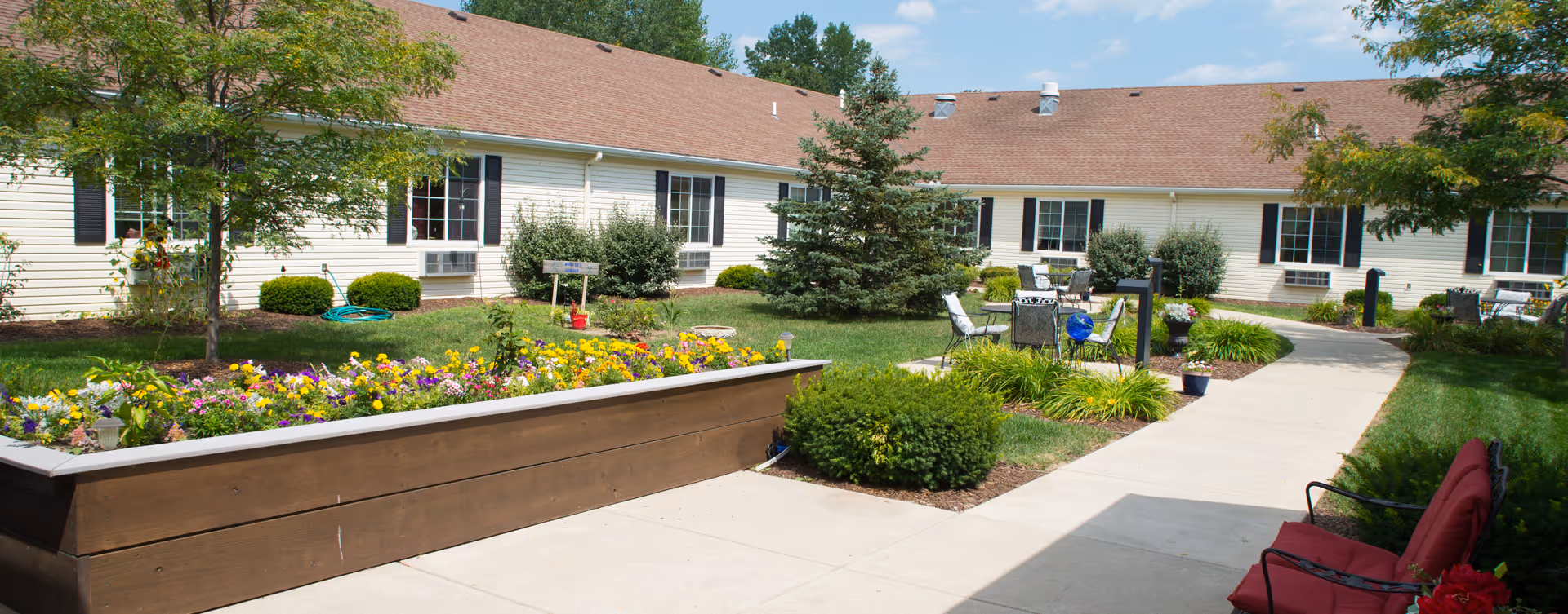 Outdoor courtyard area at Bickford of Midland featuring a raised flower bed with colorful flowers, green shrubs, trees, a paved walkway, and several seating areas with chairs and tables under a clear blue sky.