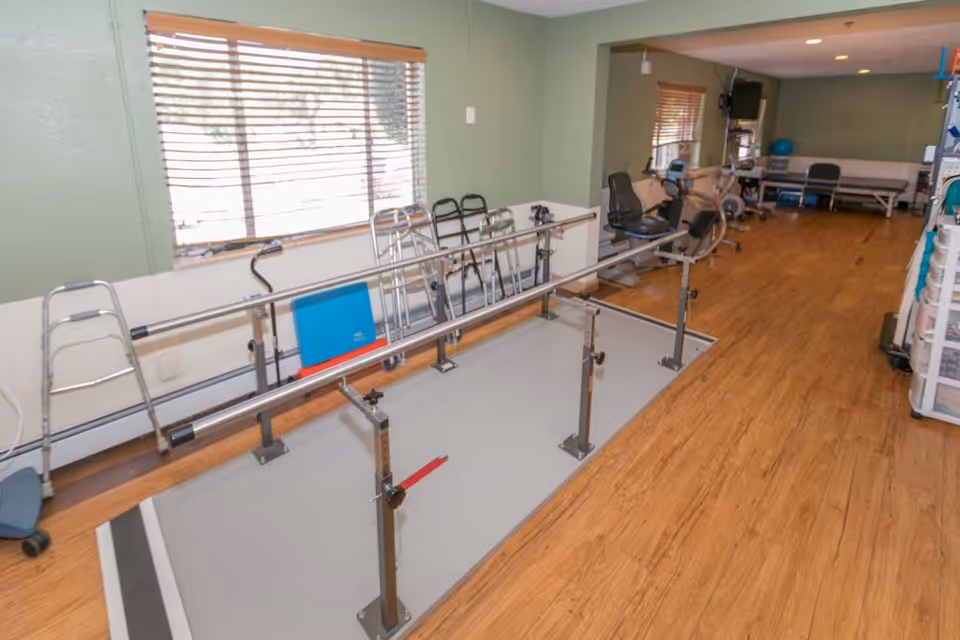 Rehabilitation exercise room with parallel walking bars, walkers, and stationary exercise equipment on a hardwood floor.