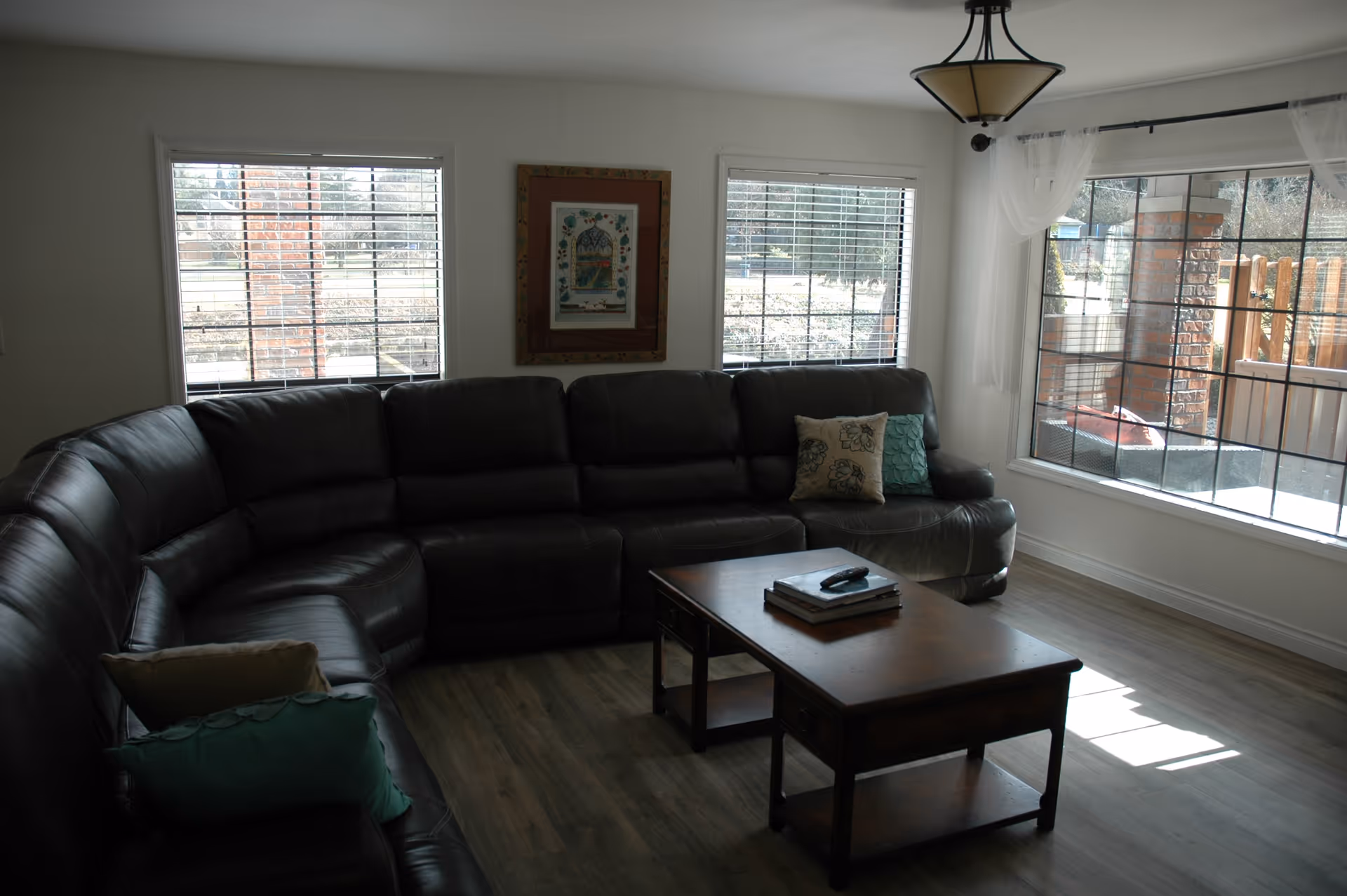 A living room with a large dark brown leather sectional sofa adorned with decorative pillows. In front of the sofa is a wooden coffee table with books and a remote control on it. The room has three large windows with white blinds, allowing natural light to enter. A framed picture hangs on the wall between two windows, and a ceiling light fixture is visible above.