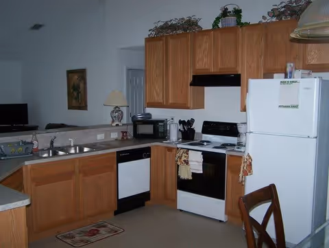 A kitchen area with wooden cabinets, a white refrigerator, a white stove with oven, a dishwasher, a microwave, a coffee maker, and a double sink. There are decorative plants on top of the cabinets and a chair partially visible in the foreground.